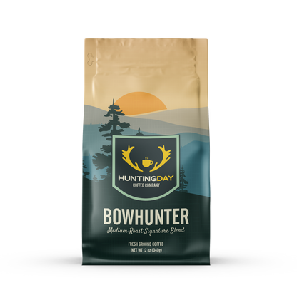A bag of Huntingday Coffee Company 'Bowhunter' medium roast coffee, featuring a landscape design with a golden sun, pine trees, and a dark teal mountain range.