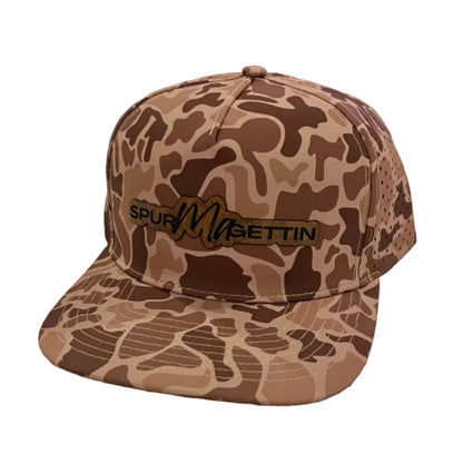 A camouflage baseball cap with a brown and tan pattern, featuring a black and gold embroidered logo that reads 'SPUR MAGETTIN'