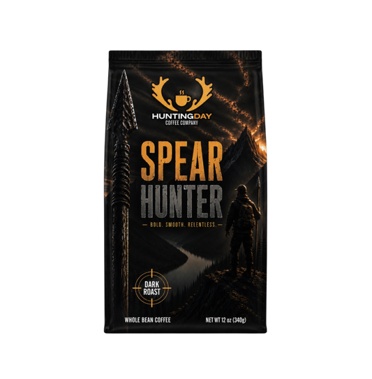 A 12 oz bag of Hunting Day Coffee Company's Spear Hunter Dark Roast whole bean coffee. The packaging is black with a dramatic graphic of a hunter standing in a dark forest at night under a fiery sky, with the product name 'SPEAR HUNTER' in large gold and gray letters.